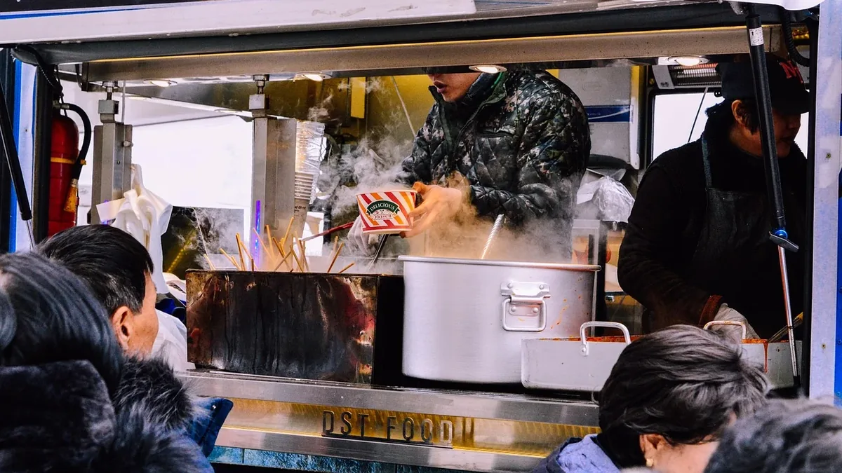 A food truck is visible, with a man in a black jacket standing behind a large pot, serving food to a line of people waiting to order.