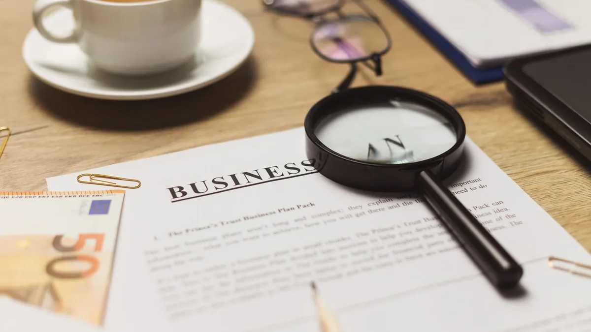 A magnifying glass on top of a business plan document, with a cup of coffee, glasses, and a 50 Euro bill in the background.
